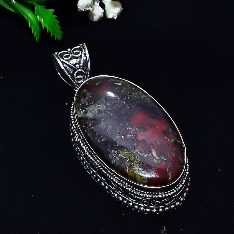 May include: Oval pendant with a polished gemstone, featuring a mix of deep red, green, and dark gray hues. The stone is set in a decorative silver-colored metal frame with intricate detailing. The pendant has a loop for a chain.