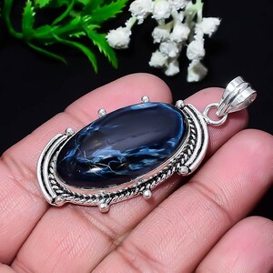 Puede incluir: Colgante ovalado con una piedra preciosa de pietersita azul pulida, que muestra vetas blancas y doradas, engastada en un marco de metal plateado. El colgante tiene una fianza decorativa. El colgante se sostiene en una mano.