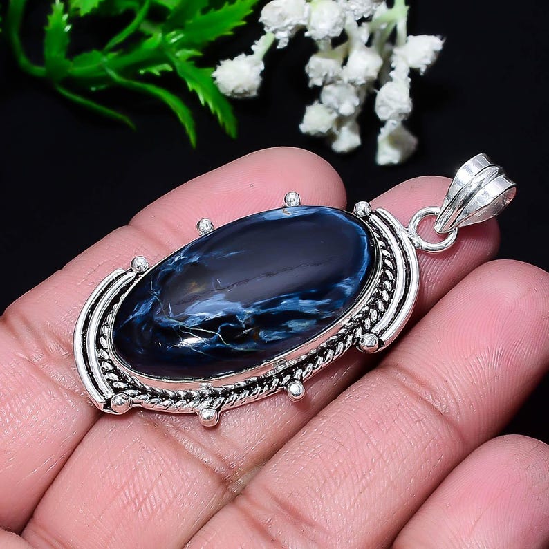 Puede incluir: Colgante ovalado con una piedra preciosa de pietersita azul pulida, que muestra vetas blancas y doradas, engastada en un marco de metal plateado. El colgante tiene una fianza decorativa. El colgante se sostiene en una mano.