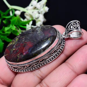 May include: Oval pendant with a polished, multi-colored stone in a silver setting. The stone features a mix of dark red, green, and black hues. The silver setting has intricate detailing around the stone and a decorative bail.