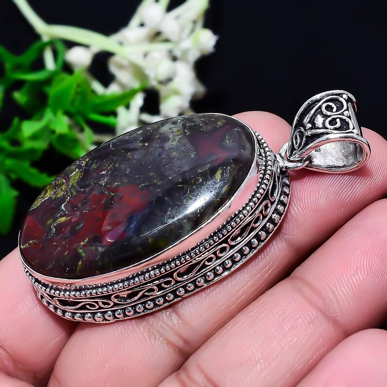 May include: Oval pendant with a polished, multi-colored stone in a silver setting. The stone features a mix of dark red, green, and black hues. The silver setting has intricate detailing around the stone and a decorative bail.