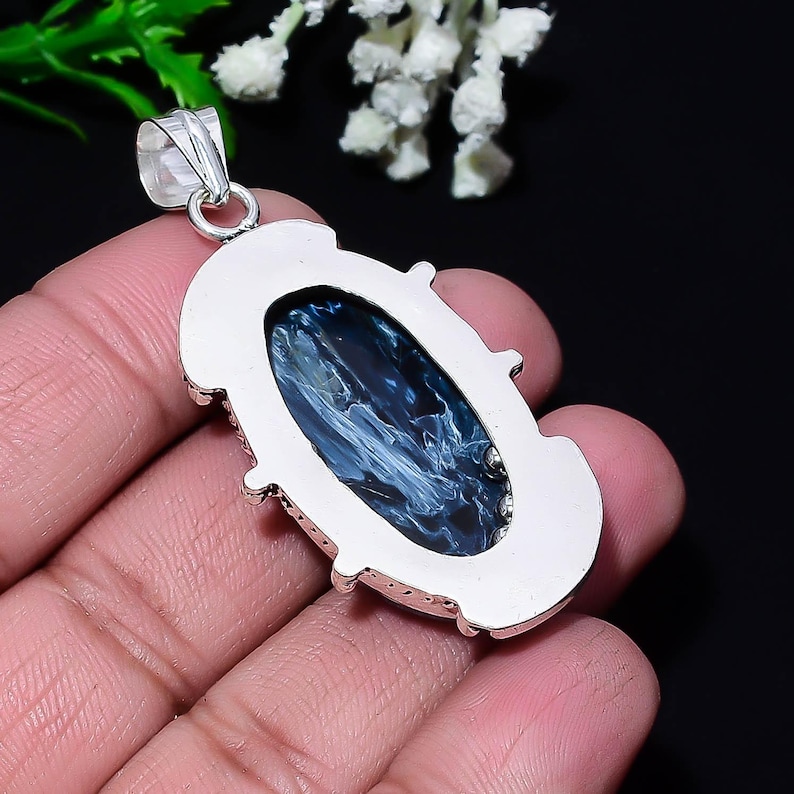 Puede incluir: Colgante ovalado con una piedra preciosa azul pulida, engastada en un marco de metal plateado. La piedra tiene un patr&oacute;n arremolinado de azul y blanco. El colgante tiene un lazo.