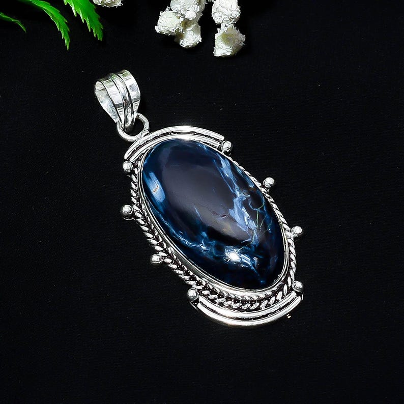 Puede incluir: Colgante ovalado con una piedra preciosa azul pulida, con vetas blancas, engastada en un marco de metal plateado. El marco tiene un borde decorativo en forma de cuerda y una argolla para una cadena. El colgante mide unos 5 cm de largo.