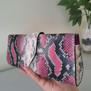 Könnte beinhalten: Eine Clutch mit rosa, schwarzen und weißen Schlangenmuster und einem Klappenverschluss. Die Clutch wird in einer Hand gehalten, wodurch das Design und die Textur des Kunstschlangenmaterials hervorgehoben werden. Die Innenseite ist einfarbig rosa.