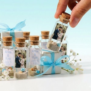 May include: A clear glass bottle with a cork stopper containing sand, small seashells, a blue bead, and a photo of a couple. A label on the bottle reads Anna & Kyle June 15, 2024. The bottle is displayed with a white gift box tied with a light blue ribbon.