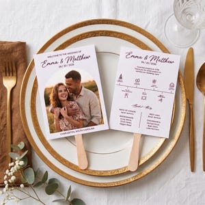 May include: Two wedding program fans with wooden handles. One fan features a photo of a couple with the text Welcome to the Wedding of Emma & Matthew 06/20/2026. The other fan displays a wedding timeline, bridal party names, and event details.