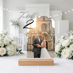 May include: A clear acrylic rectangular table sign featuring the black text "Table 2" and an arched photo of a couple in front of a stone church. The sign is supported by a rectangular light-colored wooden base, positioned on a white surface.