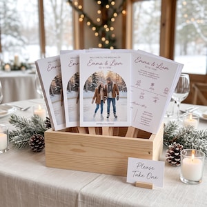 May include: A stack of wedding program fans in a light wood crate. Each fan features the text Welcome to the Wedding of Emma & Liam 12/12/2026, a photo of a couple on ice skates, and a timeline. A small sign in front reads Please Take One.