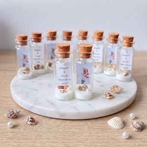 May include: A collection of small glass bottles with cork stoppers, each containing white sand, a small seashell, and a rectangular tag featuring a printed photograph of a couple and the text Megan & Oscar March 14, 2026, arranged on a round surface.