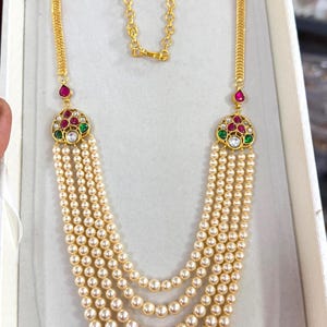Peut inclure: Un collier doré avec plusieurs rangées de perles de couleur crème. Le collier présente des pendentifs décoratifs avec des pierres précieuses rouges, vertes et blanches. La chaîne est de conception délicate.