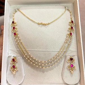May include: A gold-toned necklace and matching earrings set. The necklace features three strands of small, white pearls and is accented with red gemstones and gold detailing. The earrings mirror the necklace design, with red gemstones and pearl accents.