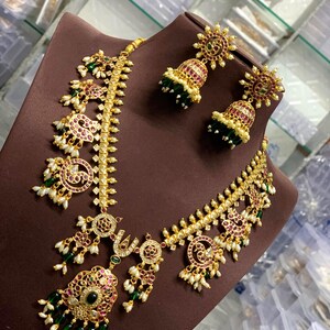 Puede incluir: Juego de collar y pendientes dorados con detalles intrincados. La joyería presenta piedras preciosas rojas y verdes, perlas blancas y un diseño floral. Los pendientes tienen forma de campana.