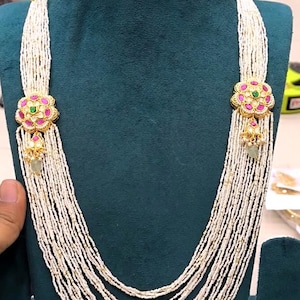 May include: A multi-strand necklace featuring numerous rows of small white seed beads, accented by two gold-toned floral motifs set with pink stones and a central green stone. Matching stud earrings feature the same floral design with dangling beads.