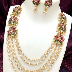 May include: A gold-toned necklace and earring set. The necklace features multiple strands of faux pearls and floral pendants with red, green, and white accents. The earrings mirror the floral design. The jewelry is displayed on a white bust.