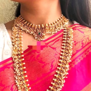 Puede incluir: Juego de joyas doradas con collar, gargantilla y pendientes. El collar y la gargantilla tienen diseños intrincados con pequeñas perlas blancas y detalles verdes. Los pendientes reflejan el diseño. Las joyas se colocan sobre una tela rosa vibrante.