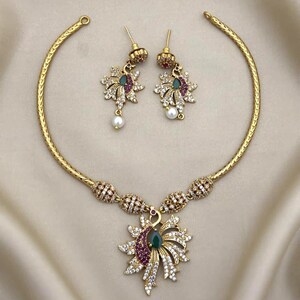 Puede incluir: Un conjunto de collar y pendientes en tono dorado con un colgante inspirado en un pavo real y pendientes a juego. La joyería está adornada con cristales blancos, pequeñas piedras rosas y una piedra verde en forma de lágrima en cada pieza.