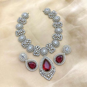 Puede incluir: Un conjunto de joyas plateadas con un collar y pendientes a juego. El collar y los pendientes tienen un diseño floral con piedras redondas y transparentes. Los pendientes y el collar presentan grandes piedras preciosas rojas en forma de lágrima. El conjunto de joyas se exhibe sobre una tela beige.