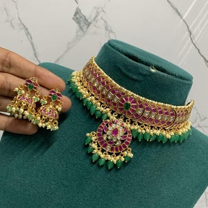 May include: A gold-toned choker necklace and matching jhumka earrings set featuring magenta-colored circular inlays, clear stones, and dangling green beads. The necklace includes a central circular pendant with a floral-inspired design.