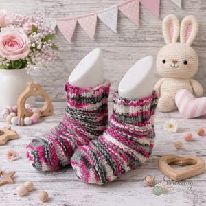 Könnte beinhalten: Ein Paar handgestrickte Babysocken in Rosa, Grau und Weiß. Die Socken werden auf weißen Formen präsentiert, mit einem Holzhintergrund und einem gehäkelten Hasen im Hintergrund. Die Socken haben ein geripptes Bündchen und ein gestreiftes Muster.
