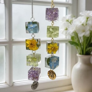 Puede incluir: Dos adornos colgantes decorativos, cada uno compuesto por cubos de resina transparente con flores incrustadas de varios colores. Las flores incluyen flores amarillas, azules y moradas. Cada adorno tiene una cadena de metal y un dije con la cara y el nombre de una mascota.