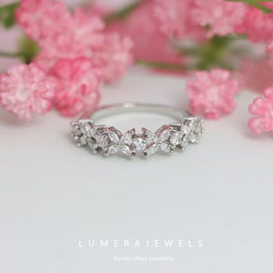 May include: A silver ring with a floral design, featuring multiple clear gemstones. The ring is set against a soft white background with pink flowers in the background. The text "LUMERA JEWELS" and "Handcrafted Jewellery" are at the bottom.