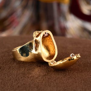 Op de afbeelding: Een goudkleurige ring met een open compartiment. De ring heeft een gedetailleerd ontwerp en een scharnierende opening, die een binnenruimte onthult. De ring wordt gepresenteerd op een bruin oppervlak, dat het ingewikkelde vakmanschap en ontwerp laat zien.