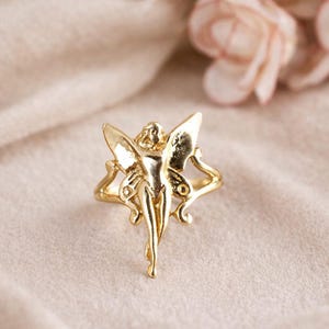 May include: A gold-colored ring featuring a fairy design. The fairy has outstretched wings and is centered on the ring's band. The ring is set against a soft, neutral background, highlighting the intricate details of the jewelry.