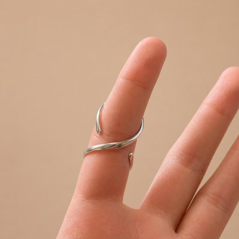K&ouml;nnte beinhalten: Ein silberfarbener Ring mit einem einzigartigen, flie&szlig;enden Design. Der Ring umschlie&szlig;t den Finger mit zwei offenen Enden und erzeugt einen modernen und eleganten Look. Der Ring wird auf einem Finger vor einem neutralen Hintergrund getragen.