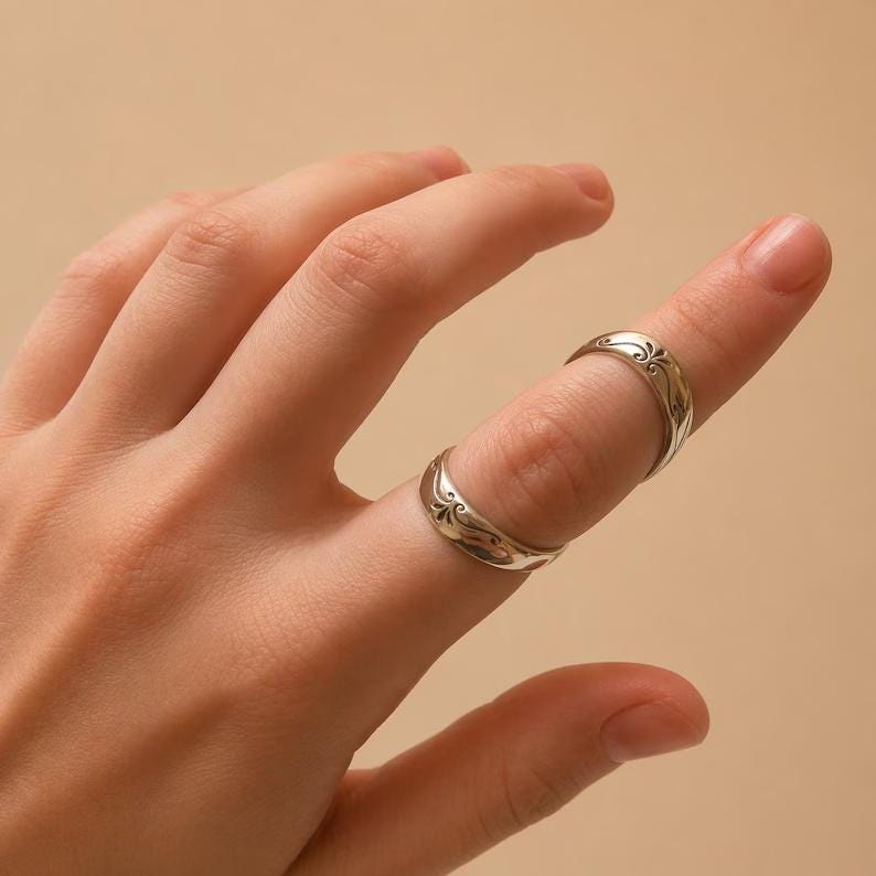 K&ouml;nnte beinhalten: Zwei silberne Ringe mit einem eingravierten floralen Design. Die Ringe werden an einem Finger getragen und zeigen die komplizierten Details des Schmucks. Die Ringe haben eine polierte Oberfl&auml;che, die das Licht reflektiert.