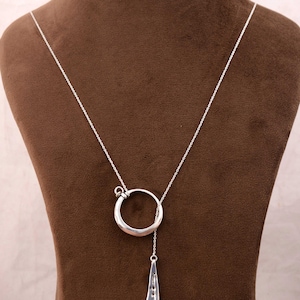 Puede incluir: Un collar plateado con un colgante circular y un dije en forma de lágrima. El collar se exhibe en un maniquí marrón, resaltando su diseño. El colgante tiene un diseño moderno.