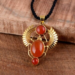 Puede incluir: Collar con colgante dorado con diseño de escarabajo, con tres piedras preciosas de color rojo anaranjado. El colgante está suspendido de un cordón negro. El collar está sobre un fondo de madera.
