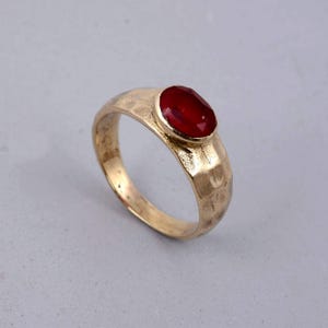 May include: A gold-colored ring with a hammered texture and a large, oval, red gemstone. The ring's band has a textured surface, and the gemstone is set in a simple bezel setting. The ring is displayed on a light gray background.