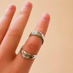 K&ouml;nnte beinhalten: Zwei silberne Ringe mit einem dekorativen floralen Design werden an einem Finger getragen. Die Ringe haben eine gebogene Form und eine polierte Oberfl&auml;che. Der Hintergrund ist hellpfirsichfarben.