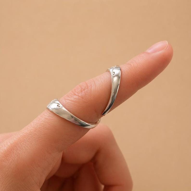 K&ouml;nnte beinhalten: Ein silberfarbener Ring wird an einem Finger getragen. Der Ring hat ein einzigartiges Design mit zwei gebogenen B&auml;ndern, die den Finger umschlie&szlig;en. Der Ring besteht aus gl&auml;nzendem Metall und hat eine glatte Oberfl&auml;che. Der Ring ist ein Schmuckst&uuml;ck.