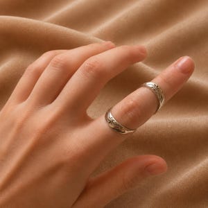 May include: Two silver rings with ornate floral designs are worn on a finger. The rings have a slightly curved shape and a polished finish. The finger is positioned against a neutral-toned background.