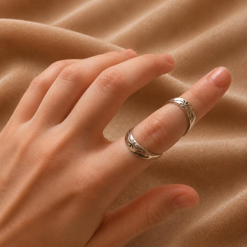 K&ouml;nnte beinhalten: Zwei silberne Ringe mit kunstvollen floralen Designs werden an einem Finger getragen. Die Ringe haben eine leicht gebogene Form und eine polierte Oberfl&auml;che. Der Finger ist vor einem neutralfarbenen Hintergrund positioniert.