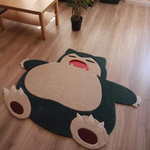 May include: A plush, character-shaped area rug featuring a dark teal body with a large, light beige belly. The character has a sleeping expression with a red mouth, white teeth, and light beige feet with dark brown circular pads and white claws.