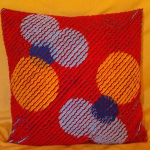 Könnte beinhalten: Ein quadratisches rotes Kissen mit strukturierter Oberfläche. Das Kissen zeigt große, kreisförmige Muster in Orange, Blau und Hellblau. Die Kreise bestehen aus parallelen Linien. Das Kissen liegt auf einer gelben Oberfläche.