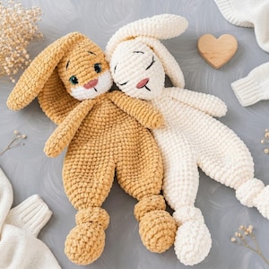 May include: Two handmade crochet bunny dolls in a warm, neutral color palette. One bunny is a light tan color, the other is white. Both have long ears, closed eyes, and are hugging each other. A wooden heart and a white sweater are in the background.