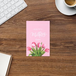 Puede incluir: Una tarjeta rosa del Día de la Madre con las palabras "Happy Mother's Day" en una elegante escritura. La tarjeta presenta un ramo de tulipanes rosas con hojas verdes. La tarjeta está sobre una superficie de madera.