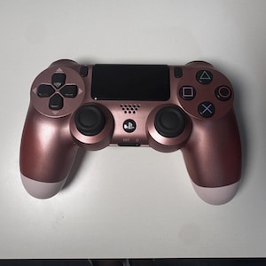 May include: A rose gold PlayStation 4 controller with black buttons and a black touchpad. The controller has a matte finish with white accents on the handles. The directional pad, buttons, and analog sticks are black.