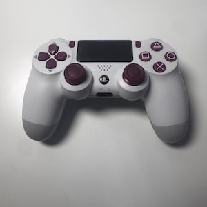 Puede incluir: Un mando blanco y gris de PlayStation 4 con detalles burdeos. El mando tiene un panel táctil, una cruceta y botones de acción. Los joysticks y botones son burdeos. El mando tiene un logotipo de PlayStation.