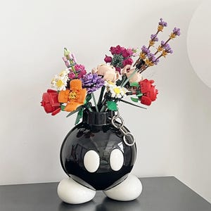 May include: A black ceramic vase shaped like a cartoon bomb, with white oval eyes and feet. The vase holds a colorful arrangement of artificial flowers, including red roses, purple lavender, and orange blooms. A silver chain is attached to the vase.