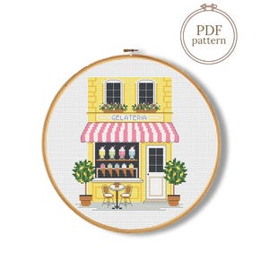 May include: A cross-stitch pattern in a wooden embroidery hoop depicting a yellow gelateria storefront with a pink and white striped awning, two potted lemon trees, a small table with two chairs, and the text GELATERIA above the window.