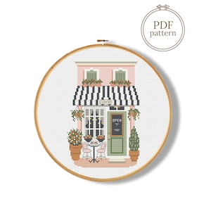 May include: Cross-stitch pattern of a pink cafe with a black and white striped awning. The cafe has a green door, small tables, and potted plants. The word "cafe" is visible on the awning. The pattern is in a wooden embroidery hoop.