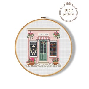 May include: A cross-stitch pattern of a craft shop, displayed in a wooden embroidery hoop. The shop features a pink facade, a striped awning, and a green door. The text "CRAFT SHOP" is above the door, and "OPEN" is on the door.
