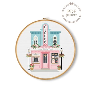 May include: A cross-stitch pattern in a wooden embroidery hoop depicting a cinema building with a light blue upper level and a pink ground floor. The design features the text CINEMA, NOW SHOWING, and TICKETS, along with street lamps and floral accents.