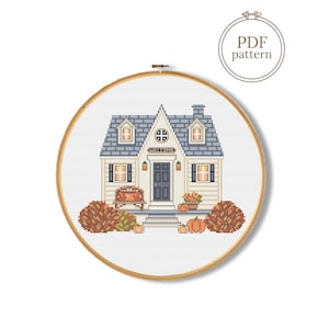 May include: A cross stitch pattern in a wooden embroidery hoop featuring a house with a blue roof, a dark door, and a welcome sign. The scene includes a bench, orange pumpkins, and autumn-colored bushes. A circular badge in the corner reads PDF pattern.