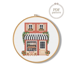 May include: A cross stitch pattern in a wooden embroidery hoop depicting a red brick building with a black and white striped awning. The storefront sign reads TAPAS AND WINE BAR, featuring shelves of bottles, glasses, and a small table with chairs.