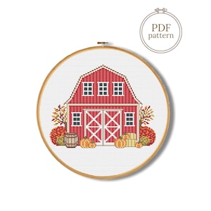 May include: A cross stitch pattern featuring a red barn with white trim, windows, and double doors. The scene includes autumn trees, pumpkins, and a wooden barrel on a white grid background, all framed within a circular wooden embroidery hoop.